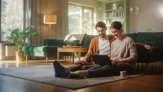 A couple sitting on the floor of a living room with contemporary decor, and using a laptop