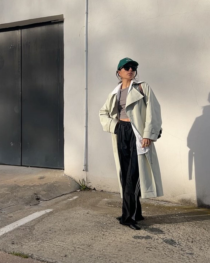 fashion influencer poses outside in a green baseball cap, an oversize trench coat, cropped gray tee, black Adidas track pants, and ballet flats