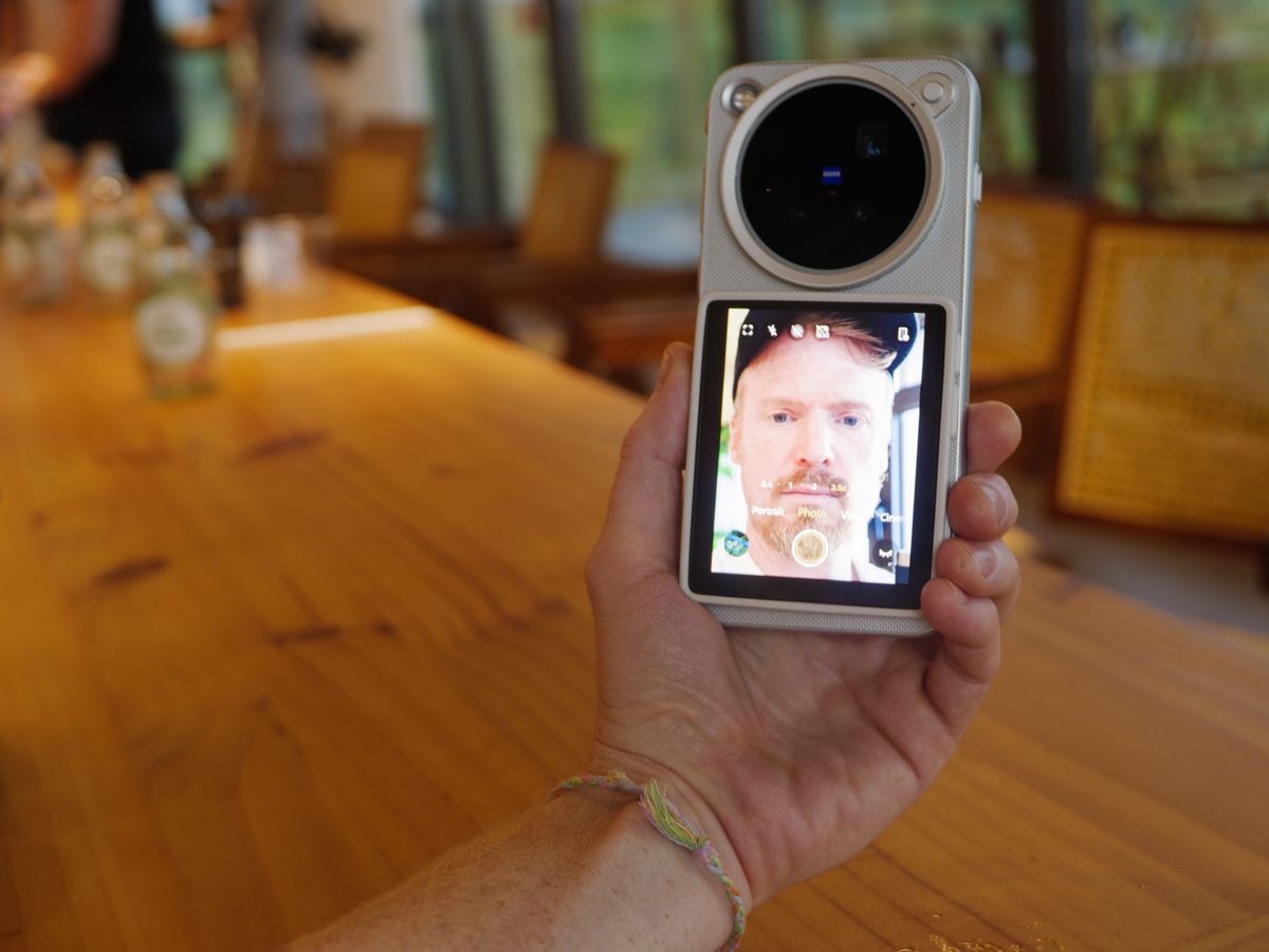 The Vivo X500 Pro Max phone in user's hand, in its dedicated case for magnetic accessories, with Vivo's selfie screen attached, and person is vlogging inside a cafe