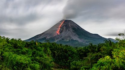 A volcano with lava running down its side. The volcano is surrounded by lush forest.