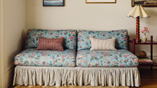 a blue floral printed couch with a skirt and two pillows. A side table holding books, a small vase, and a lamp with a twisted wood base
