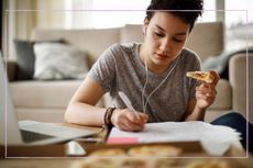 A teenage girl studying with earphones in while eating pizza