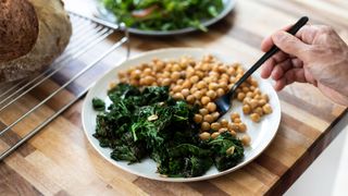 An old hand holding a fork, eating a plate of leafy green vegetables and chickpeas