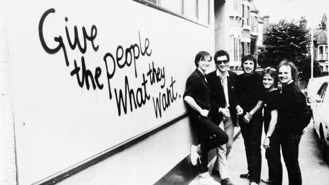 The Kinks standing next to a Give The People What They Want billboard
