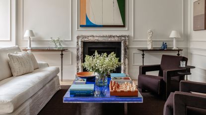 Image of a traditional white living room with a white sofa, two plum accent chairs, and a royal blue coffee table with books, flowers, and a chess set on it.