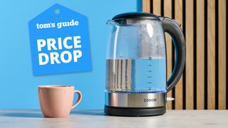 The Cosori Electric Glass Kettle filled with water next to a pink mug photographed in front of a blue blue background with a price drop tag.