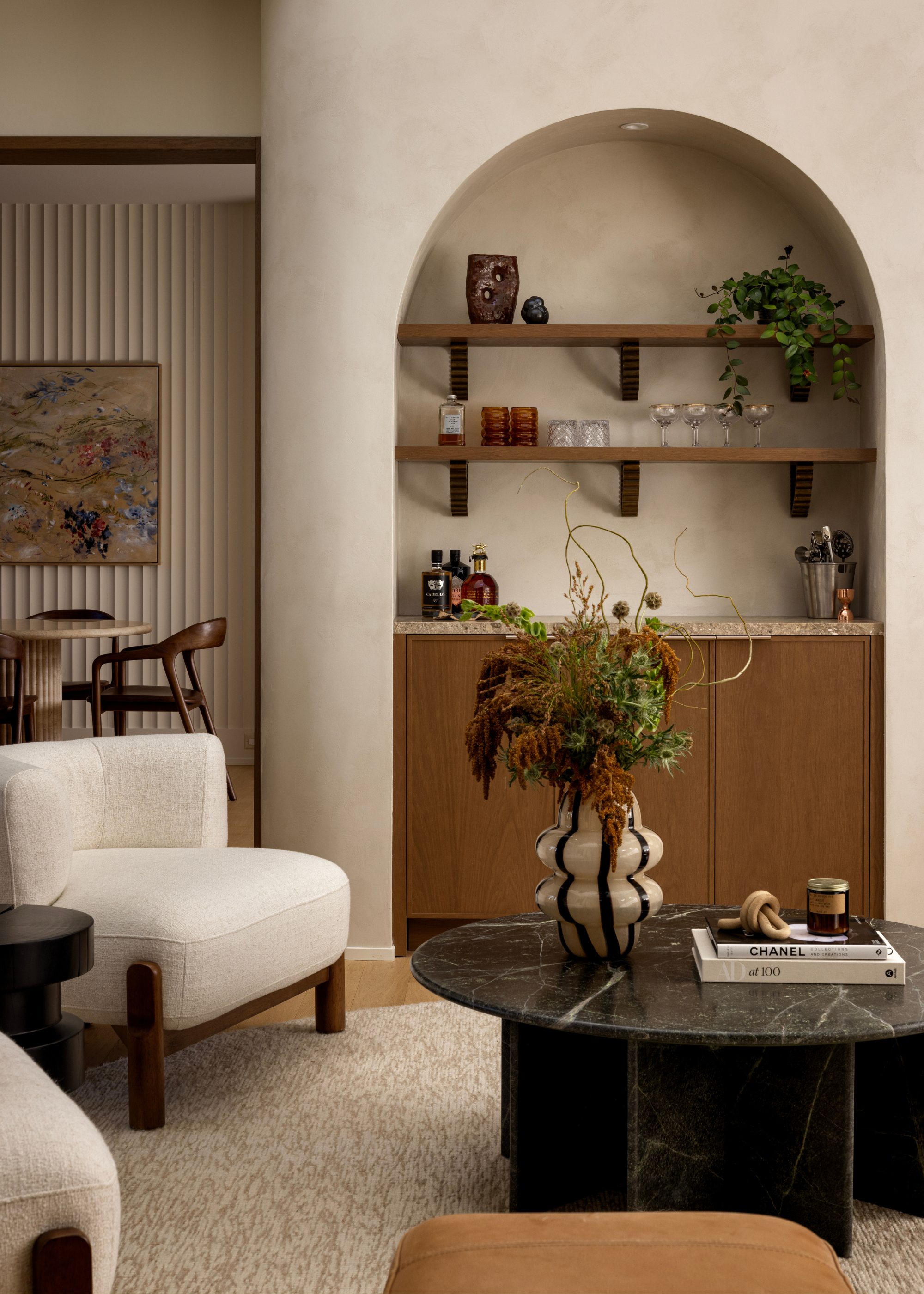 An arched bar set into the wall with floating wood shelves, a round marble, a fuzzy rug, a set of white armchairs