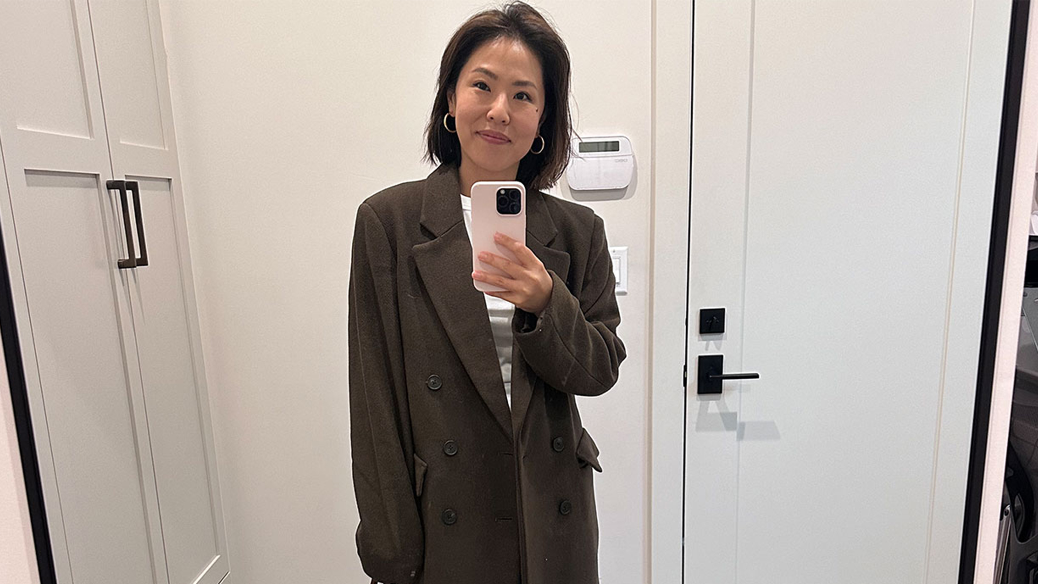 a woman stands in the mirror wearing a brown coat and a white t-shirt.
