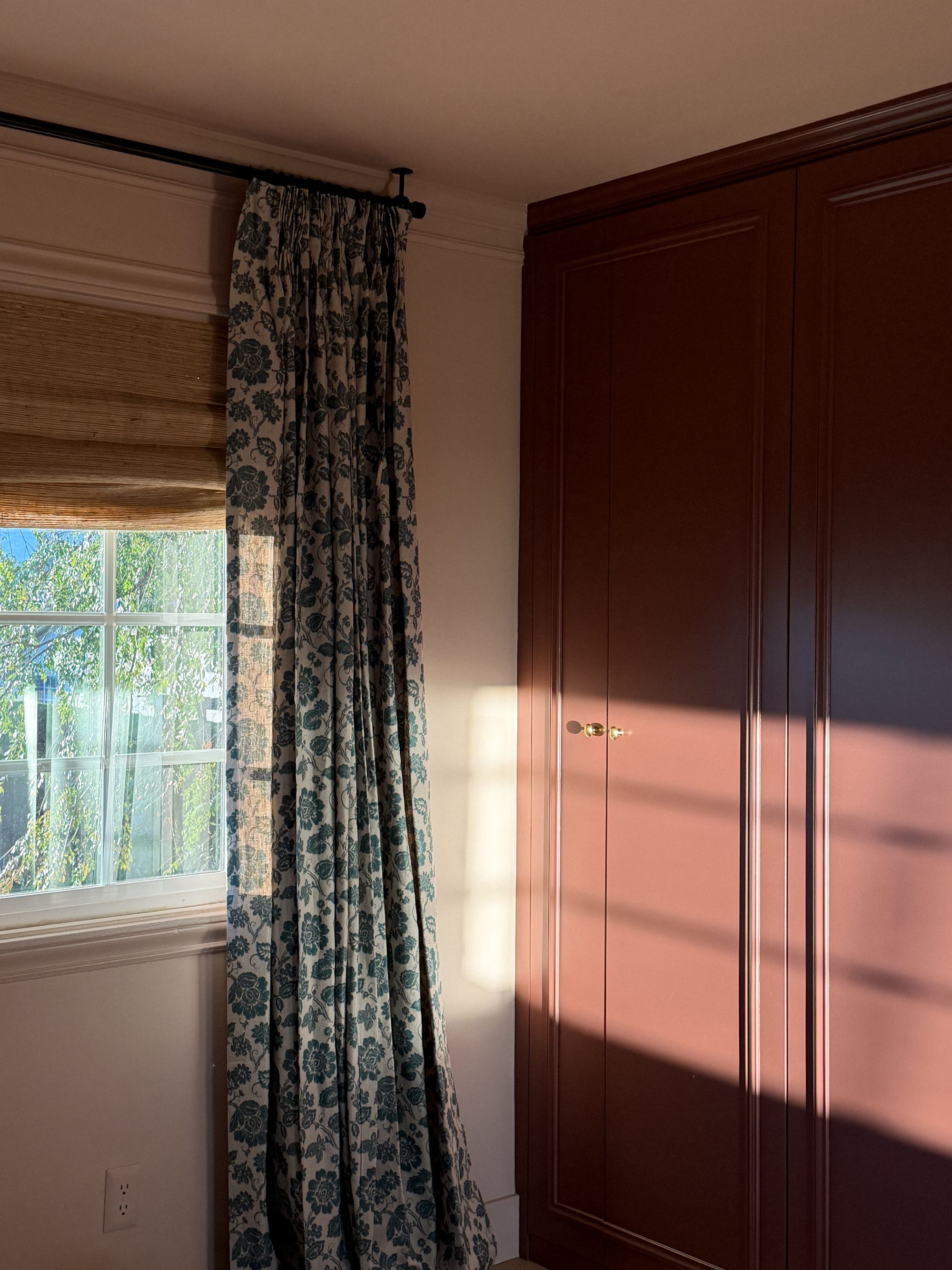 Image of the corner of a light pink room with a dark red, built-in wardrobe. There is a window on the wall with patterned curtains, and sunlight is shining across the middle of the wardrobe.