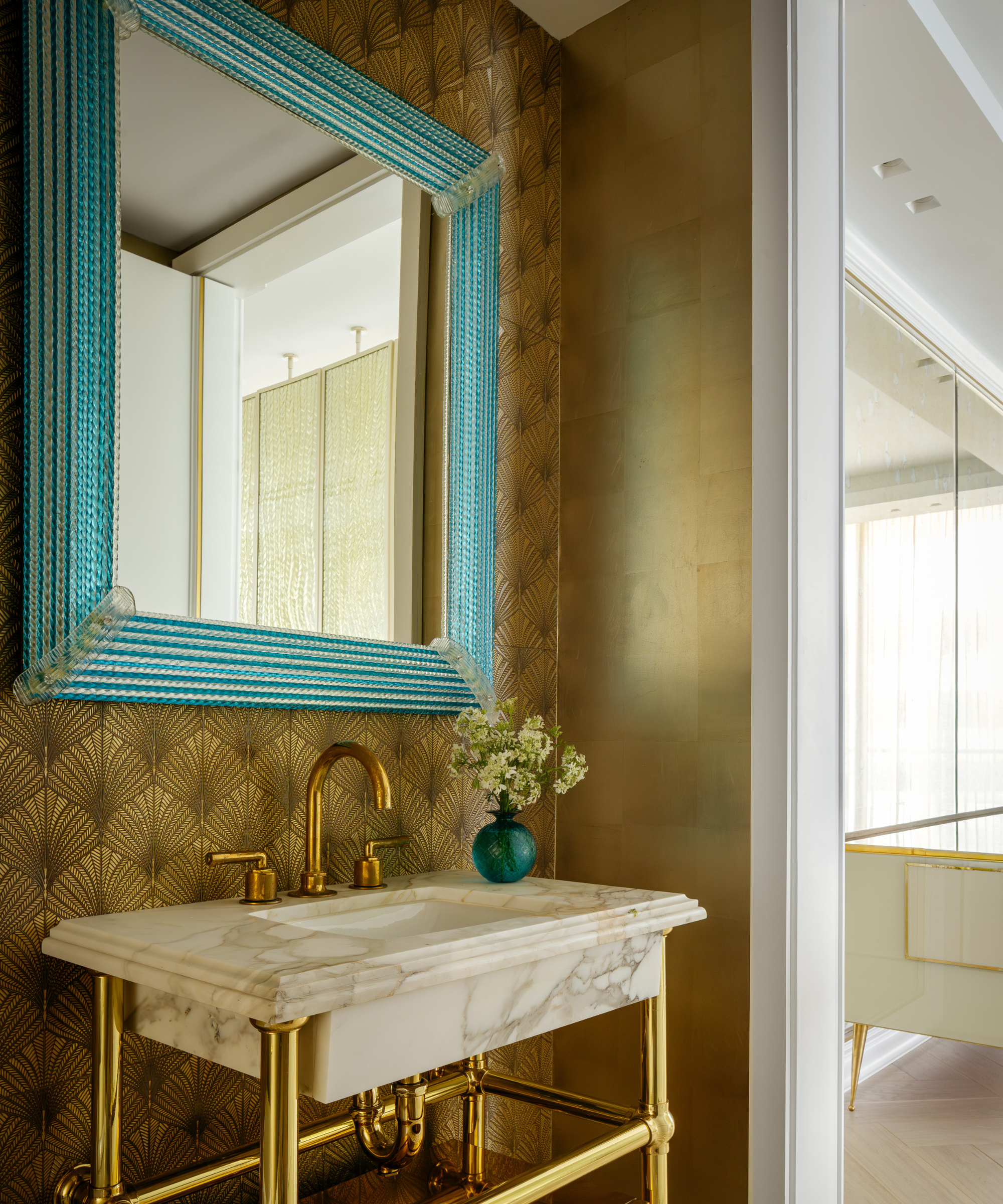 Bijou powder room featuring gold Deco-inspired wallpaper, a marble bathroom sink, and oversized teal-framed mirror overhead.
