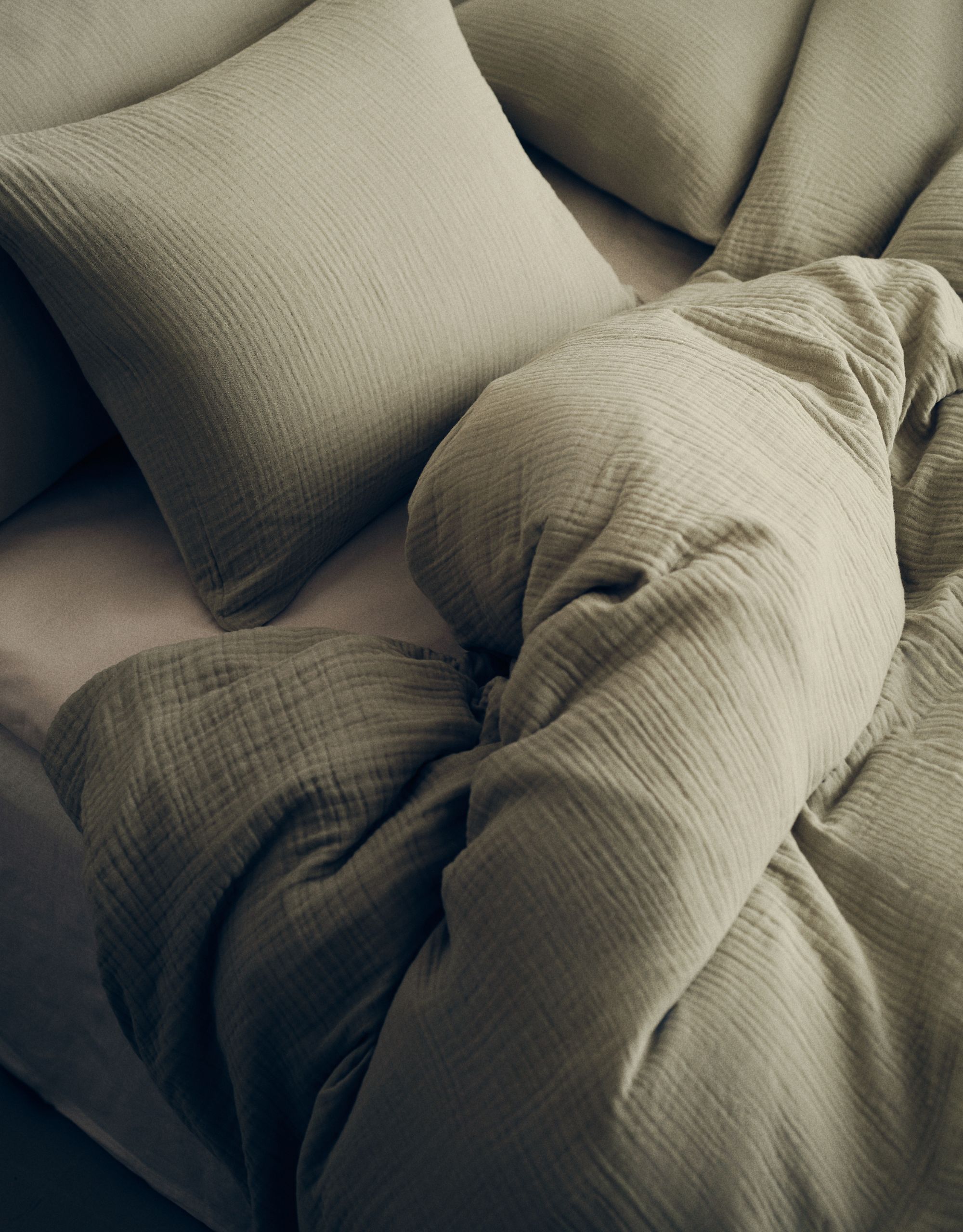 A close up of a bed with a light green muslin duvet cover and pillows against a linen sheet