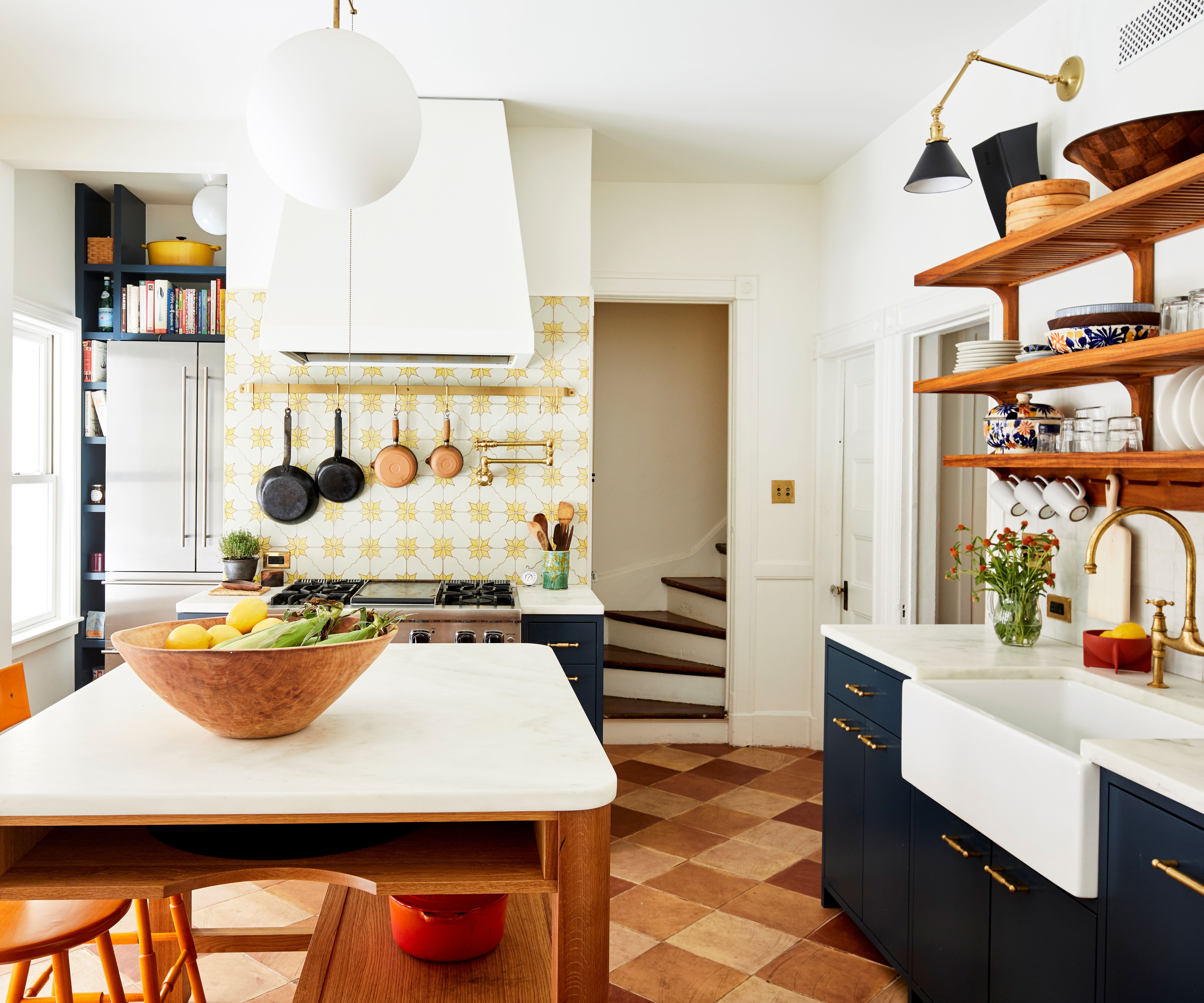 A vibrant kitchen with deep navy cabinets, a yellow patterned tile backsplash, and terracotta diamond-patterned floor tiles. Includes open wood shelving and a large white farmhouse sink