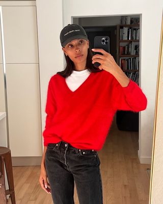 influencer Tylynn Nguyen posing for a mirror selfie wearing a red v-neck sweater layered over a white tee, baseball cap, and black jeans