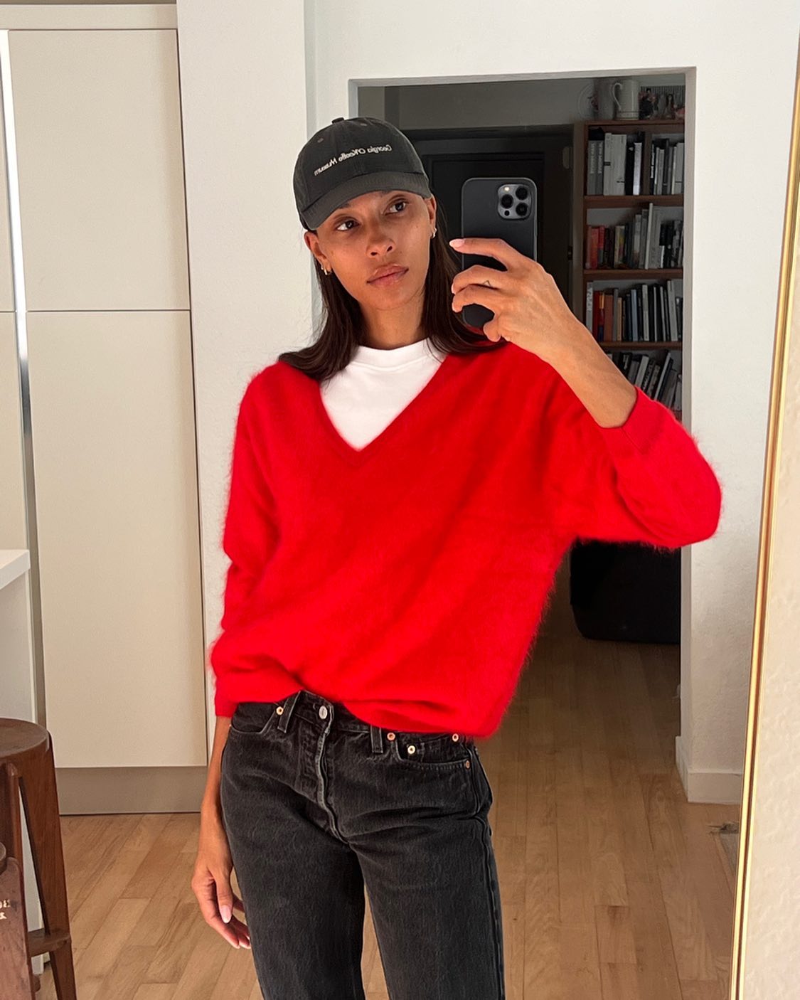 Influencer Tylynn Nguyen posing for a mirror selfie wearing a red v-neck sweater layered over a white tee, baseball cap, and black jeans.