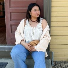 Influencer @marina_torres sits on a doorstep wearing flared jeans with ballet flats, a white lace blouse, a light cream cardigan and a red beaded necklace.