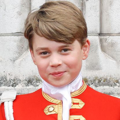 Prince George wearing a red uniform and smiling in front of a brick wall