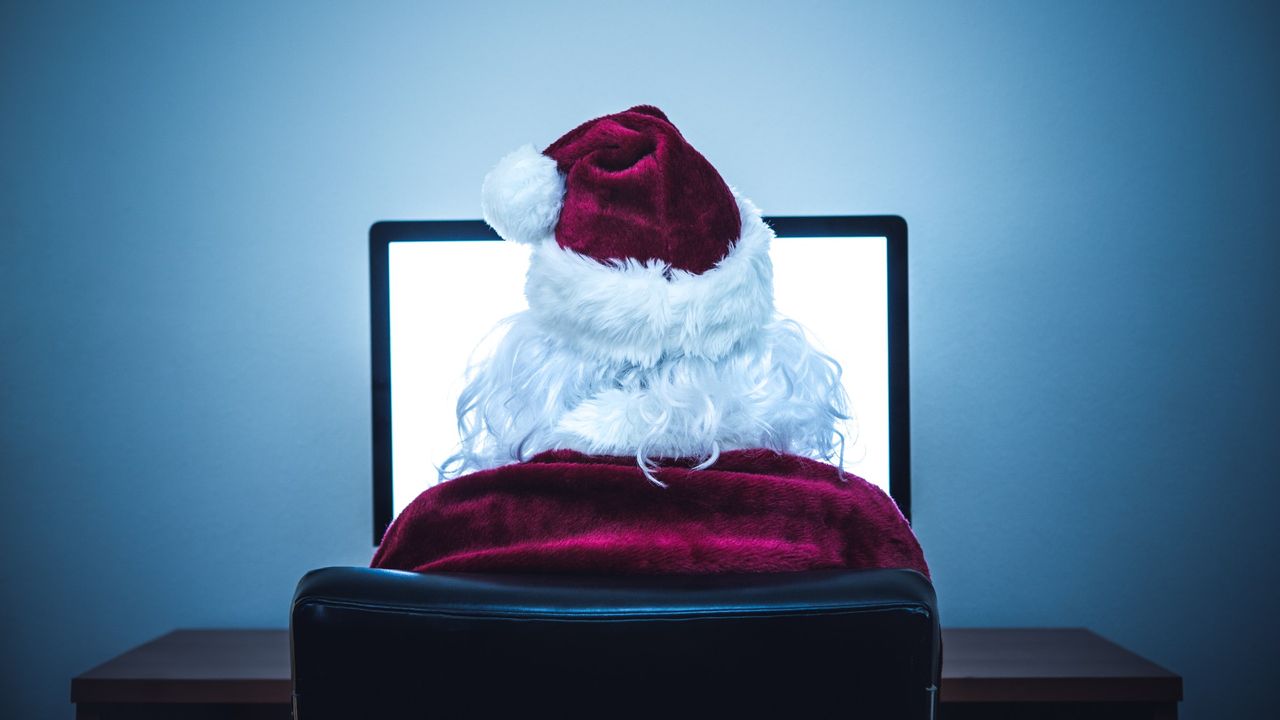 A man in a Santa Claus costume sits in front of a desktop computer