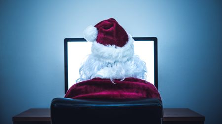 A man in a Santa Claus costume sits in front of a desktop computer