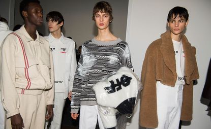 Four male models wearing looks from Off-White's collection. One model is wearing a cream tracksuit with two red stripes. Another model is wearing a white shirt with black writing and white trousers. The third model is wearing a partially faded black, white and grey striped patterned jumper, white trousers and has a black and white piece hanging over his arm that features the word "OFF". And the fourth model is wearing a white t-shirt with black writing, white trousers and a brown short fur jacket