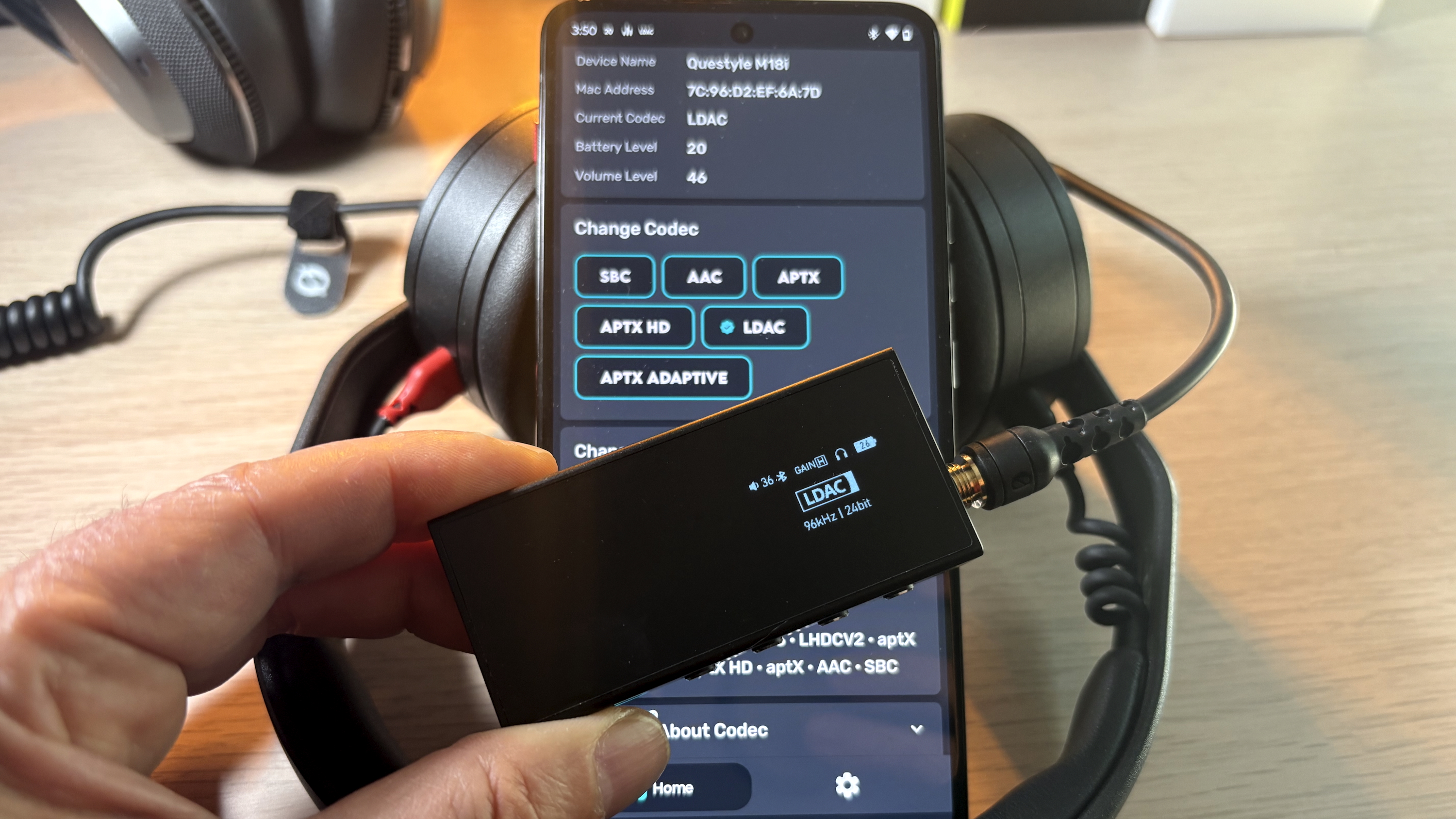 A hand holding a DAC, in front of a phone screen showing the Bluetooth codec changer app