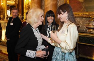 Queen Camilla wearing a black-and-white dress and speaking to actress Emily Mortimer, wearing a blue skirt and silky cream blouse and holding a glass of champagne