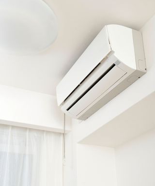 A close-up of a white ceiling corner with a rectangular white air condioning unit on the wall and white curtains underneath it