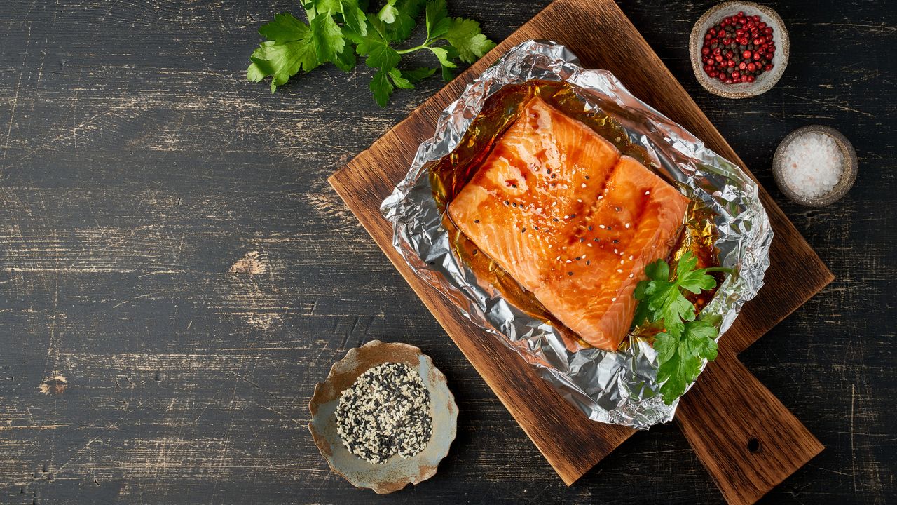 Baked salmon in a foil packet on a wooden chopping board, on a dark wooden table. There is a garnish of coriander and small bowls of seasoning.