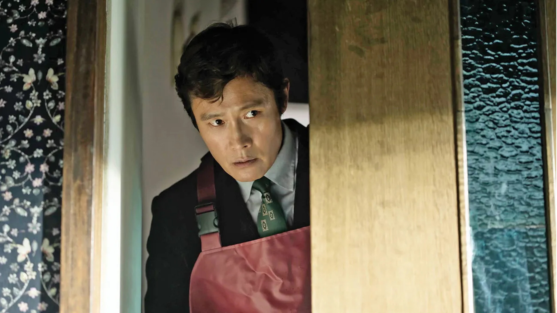 A man (Park Byung-hun as Man-su) wears an apron over a suit whole peeking through a doorway, in Park Chan-wook&#039;s 2025 movie &#039;No Other Choice.&#039;