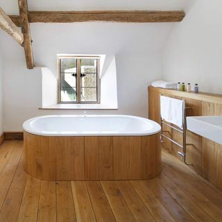Bathroom with wood panelled bath, wooden floor, wooden storage shelf along wall with a silver towel rail on it and exposed wooden beams on the ceiling
