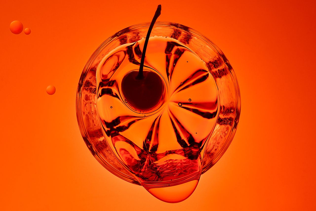 A close-up from above of a glass with liquid, featuring a cherry, showcasing swirling patterns against an vibrant orange backdrop