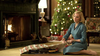 A woman in a blue dress sits on a rug in a cozy living room, wrapping a model train set near a lit fireplace and a decorated Christmas tree.