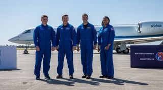 Four astronauts in their blue NASA flight suits smile while standing on an airplane tarmac with an aircraft behind them