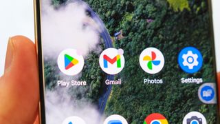 A photo of the Gmail app tile on an Android phone, with the Play Store and Google Photos apps next to it. The wallpaper is of a green landscape.