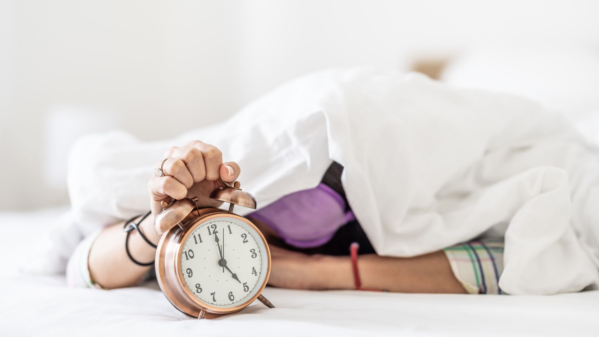 A woman under a duvet holding an alarm clock