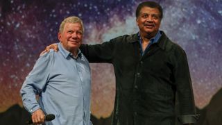 A man wearing a blue shirt stands to the left of a man wearing a black shirt, both in front of a starry background