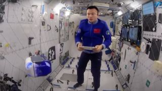 an astronaut in a blue jumpsuit tests a flying robot aboard a white-walled space station