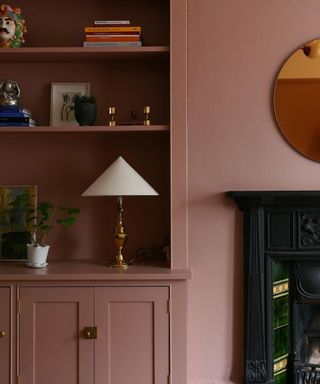 A pink living room wall with a shelf with books, lamps, and decor on it to the left and a green fireplace to the right.