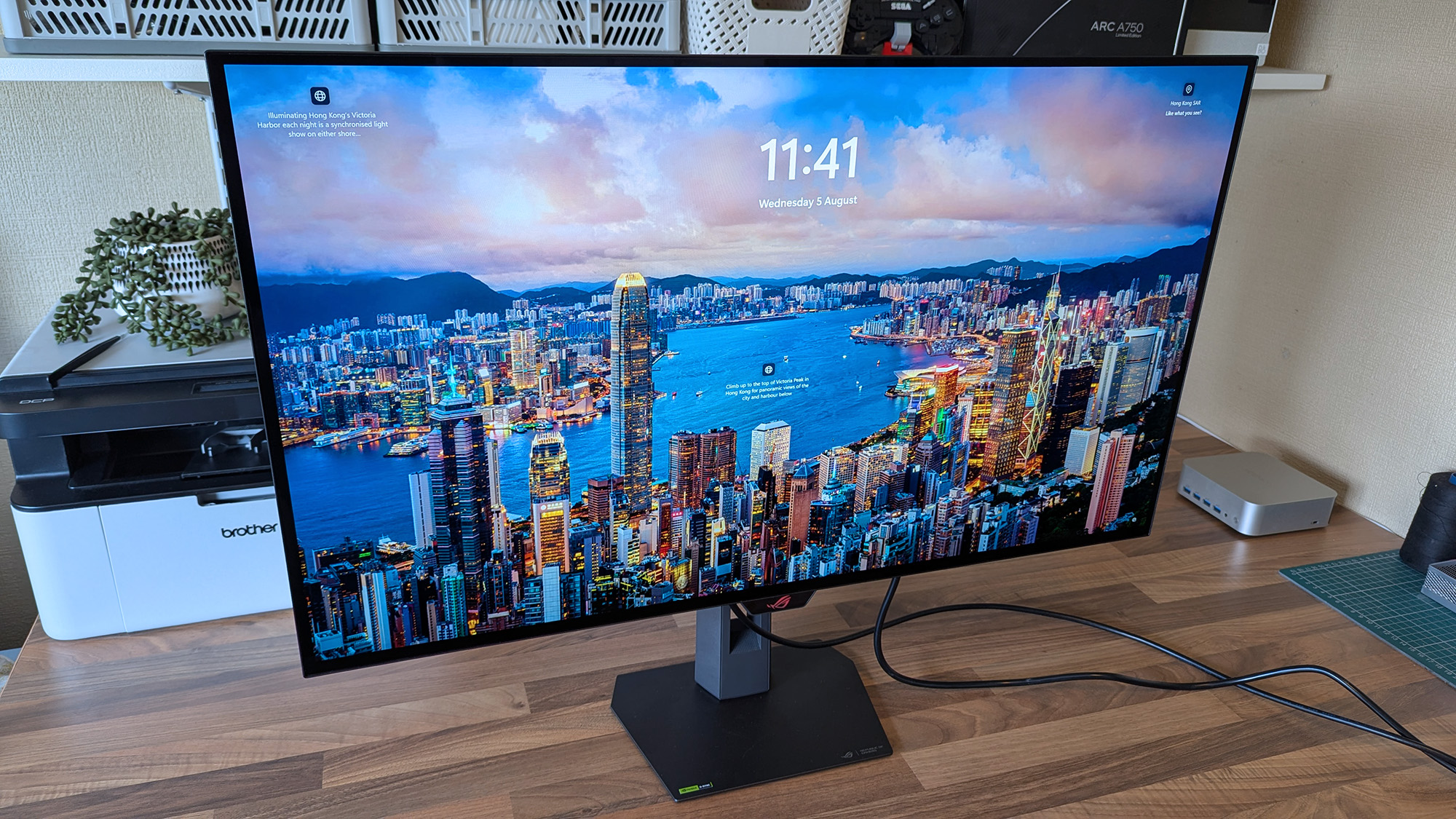 ASUS ROG Strix OLED XG32UQWMS monitor displays a vivid Hong Kong Victoria Harbor lock screen with the time, date, and descriptive text, surrounded by a desk setup.