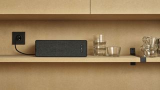 A black Sonos Ikea Symfonisk bookshelf speaker on its side, plugged into a wall socket on a set of beige shelves. Next to it is an assortment of small drinking glasses.