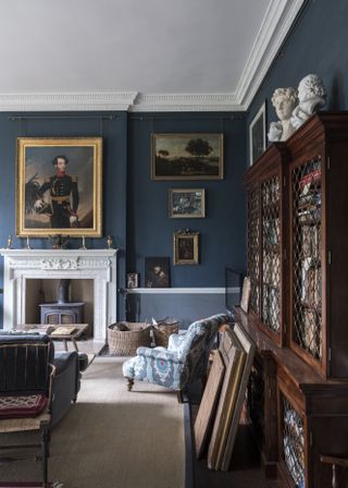 A living room painted in a dark blue color. There are multiple traditional paintings hung on the wall, a white marble fire place, and a large wooden chest filled with books on the right side of the image