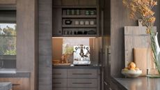 dark timber kitchen with butlers pantry with silver espresso machine, shelves above with mugs and cups, and a LED-lit backsplash. on the kitchen counter there are timber cutting boards resting upright, a bowl of fruit, and a vase filled with flowers
