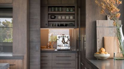 dark timber kitchen with butlers pantry with silver espresso machine, shelves above with mugs and cups, and a LED-lit backsplash. on the kitchen counter there are timber cutting boards resting upright, a bowl of fruit, and a vase filled with flowers