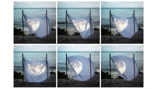 Multiple images of the moon appearing on a sheet tied to poles on a beach in front of the sea