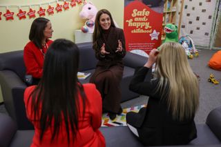 Kate Middleton wearing a plaid coat sitting on a sofa and talking to 3 women with toys in the background at a prison mother baby unit