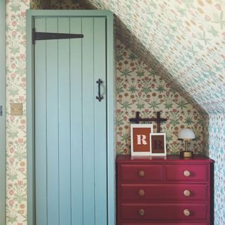 Small blue wardrobe next to a burgundy chest of drawers in front of cottage-style wallpaper