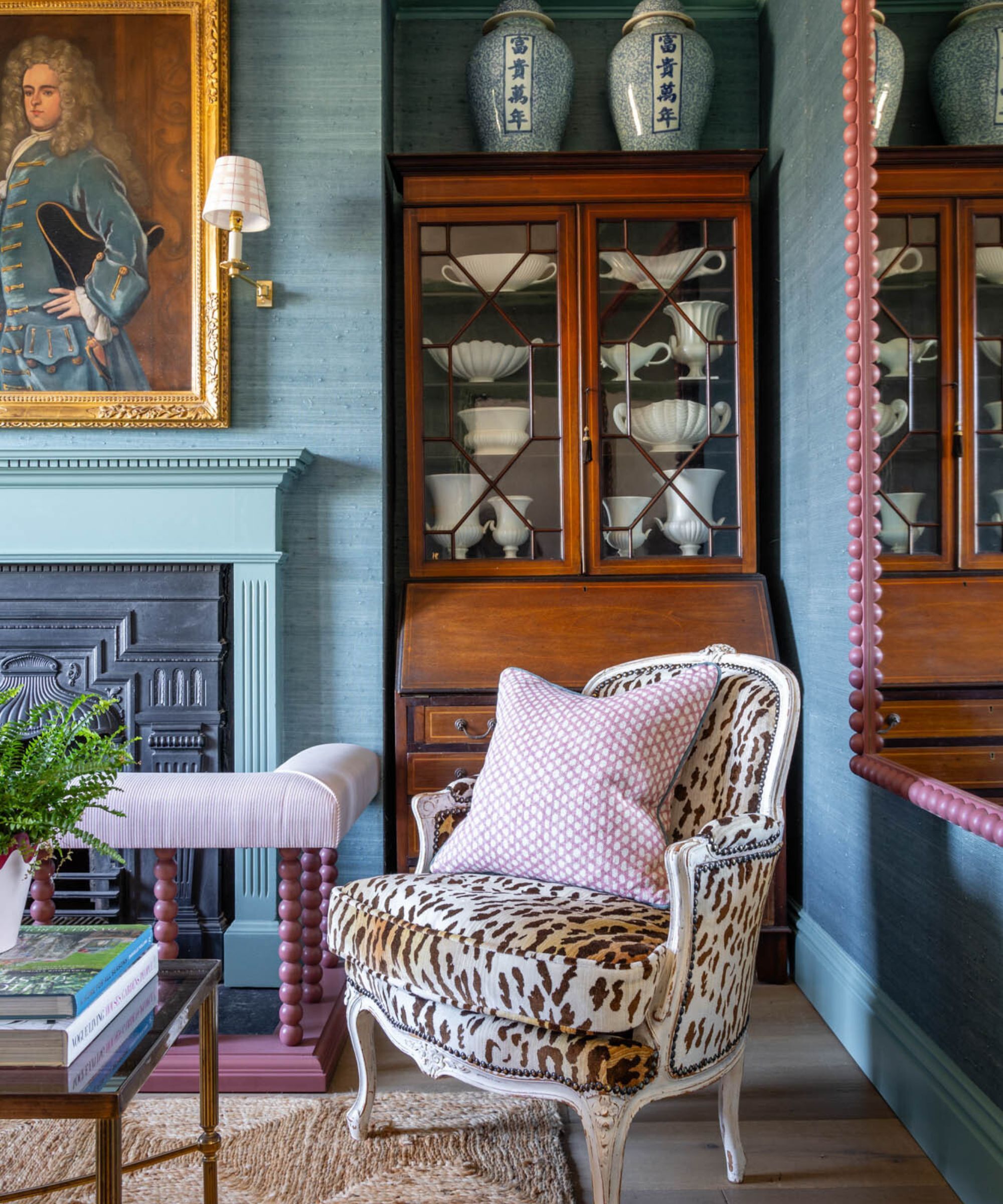 The corner of a living room. The walls have blue textured wallpaper and the skirting boards are a slightly paler shade of blue. There is a sisal rug on the floor. There is an antique chest against the wall. Infront sits a leopard print chair with a pink patterned scatter cushion. The fireplace has a large guilt painting above it with a sconce light beside it.
