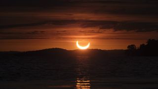 Between layers of clouds, a partial solar eclipse is seen at sunrise over Rice Lake, Ontario, as the sun, partially covered by the moon, rises in a beautiful orange sky above the trees and water.