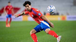 Victor Davila flicks the ball on the pitch, in his red shirt and blue shorts, ahead of the Chile vs Bolivia live stream.