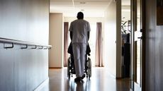 Full length rear view of male in scrubs pushing a person in on wheelchair along a corridor