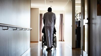 Full length rear view of male in scrubs pushing a person in on wheelchair along a corridor